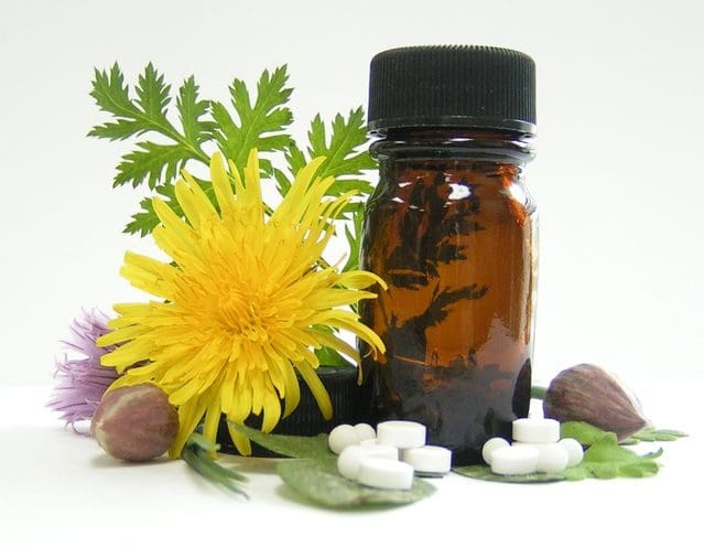 An Amber Glass Bottle With A Black Cap Surrounded By Yellow Dandelion And Purple Thistle Flowers, Green Leaves, White Tablets
